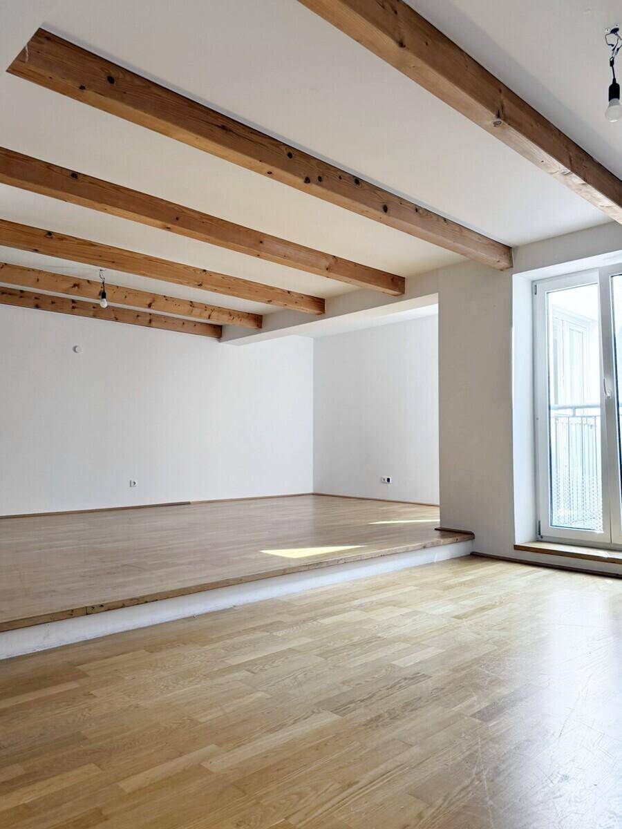 Offener Wohnbereich mit sichtbaren Holzbalken, hellem Parkettboden und Zugang zu einem französischen Balkon.