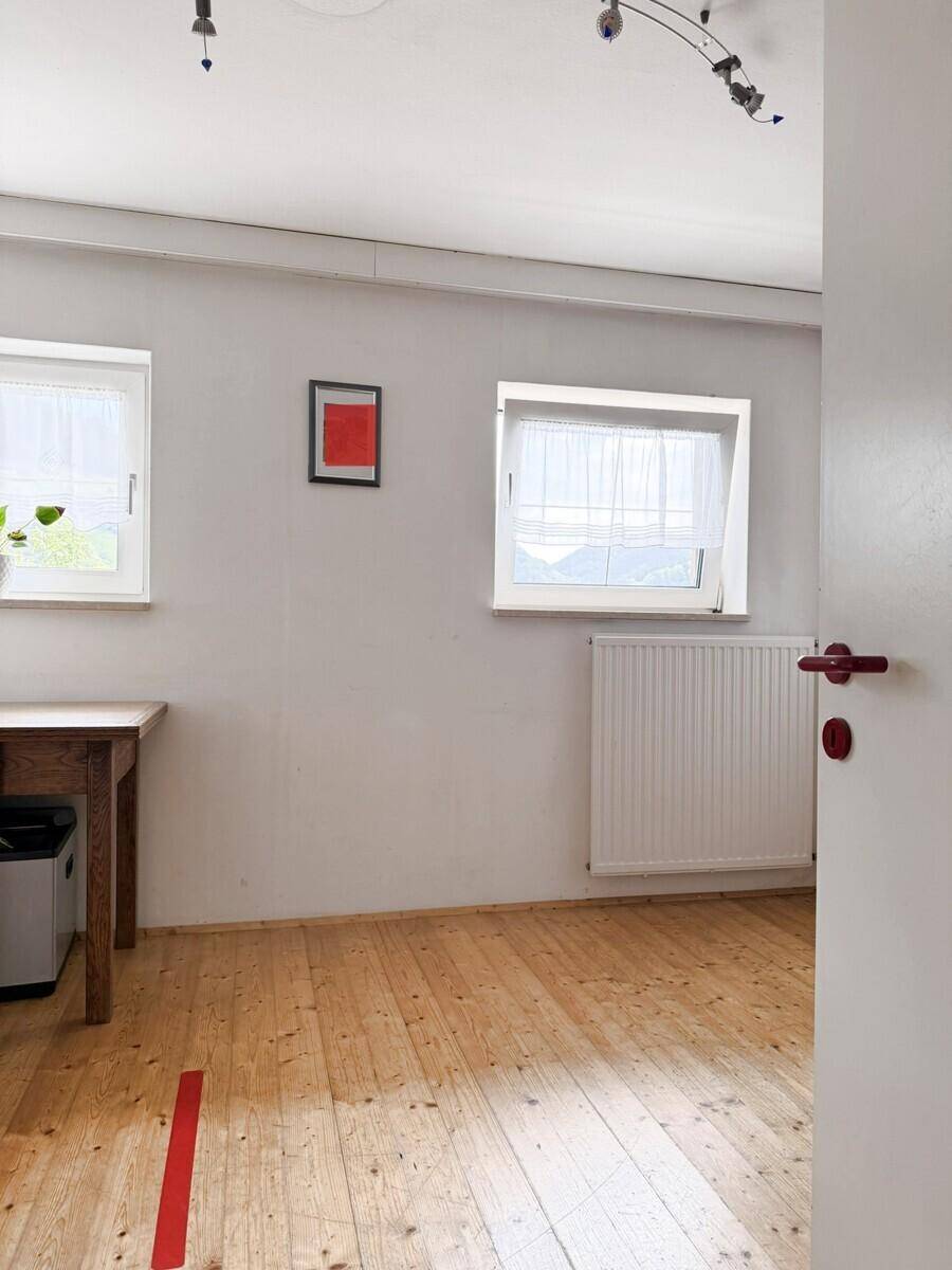 Helles Zimmer mit Holzboden, zwei Fenstern und einem Schreibtisch, ideal als Arbeitszimmer.