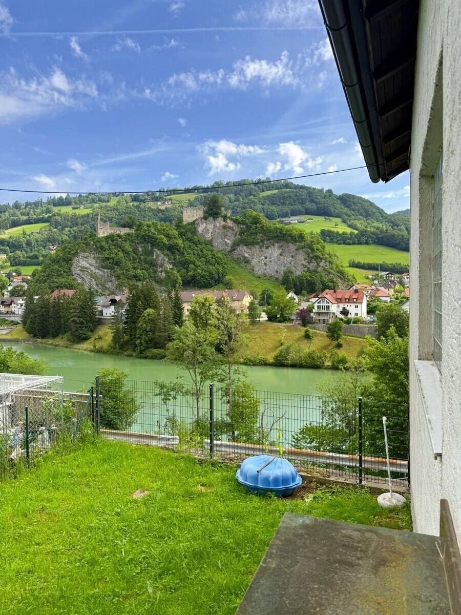 Gartenbereich mit Rasenfläche und Blick auf einen See, eine Burgruine und grüne Hügel.