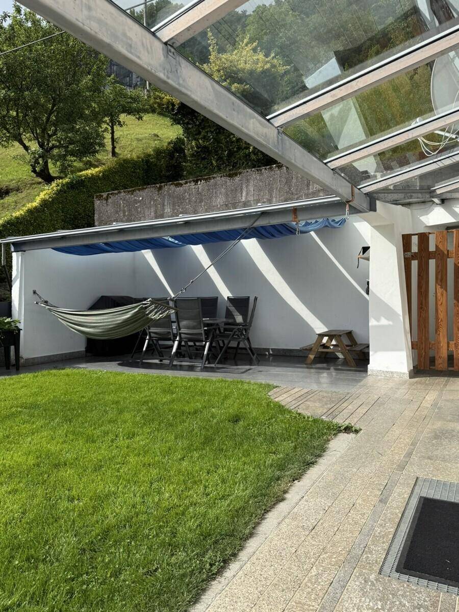 Überdachte Terrasse mit Gartenmöbeln und Hängematte, umgeben von Grünflächen und Hügeln.