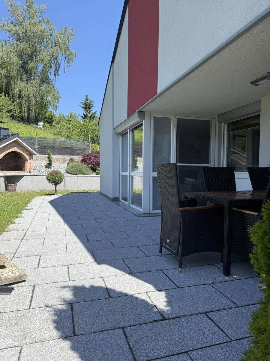 Geräumige Terrasse mit Steinplatten, die zu einer Rasenfläche und einem Pizzaofen führt.