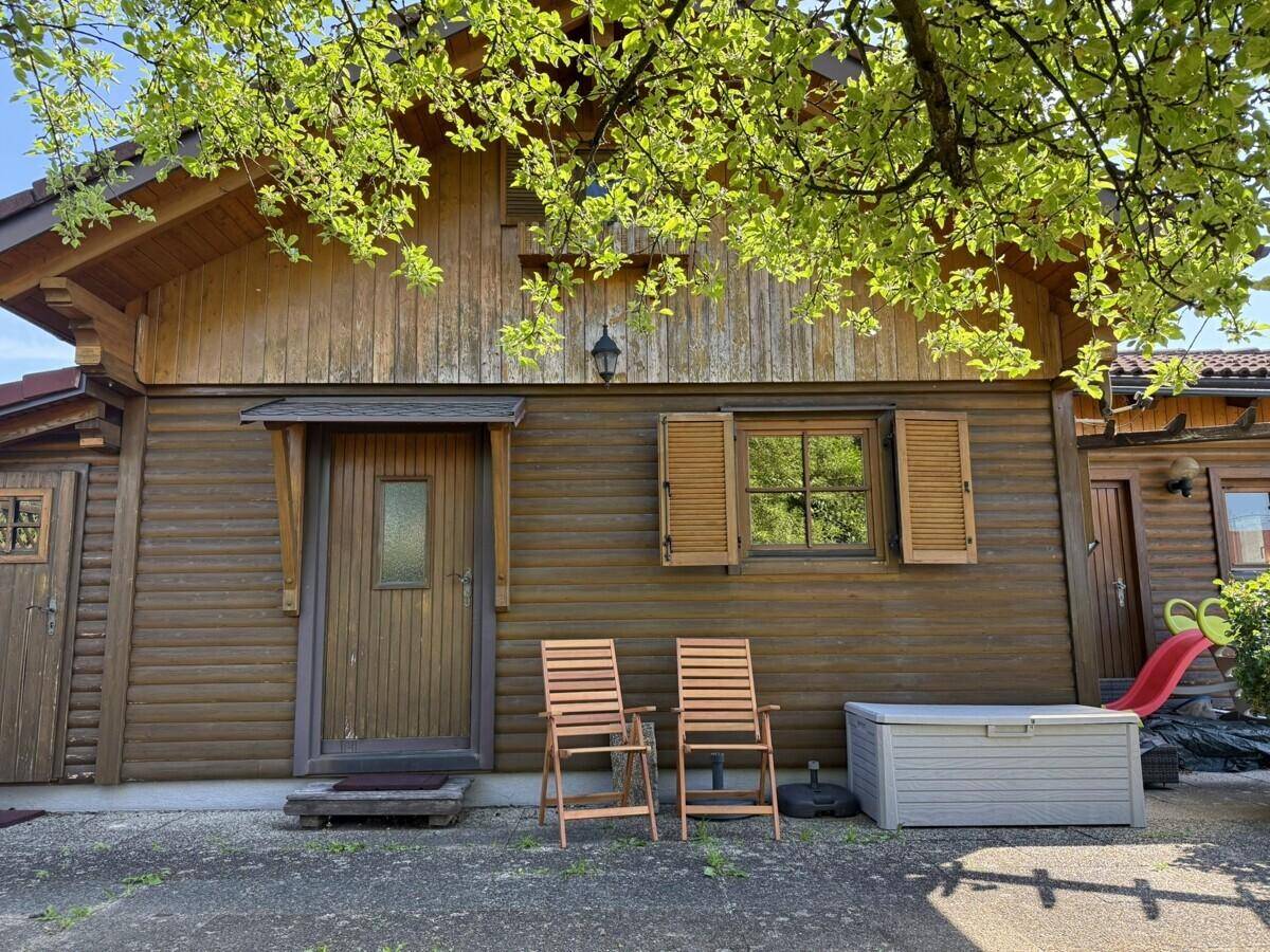 Holzhausfassade mit Fensterläden und Eingangsbereich, umgeben von Bäumen und einem Spielgerät.