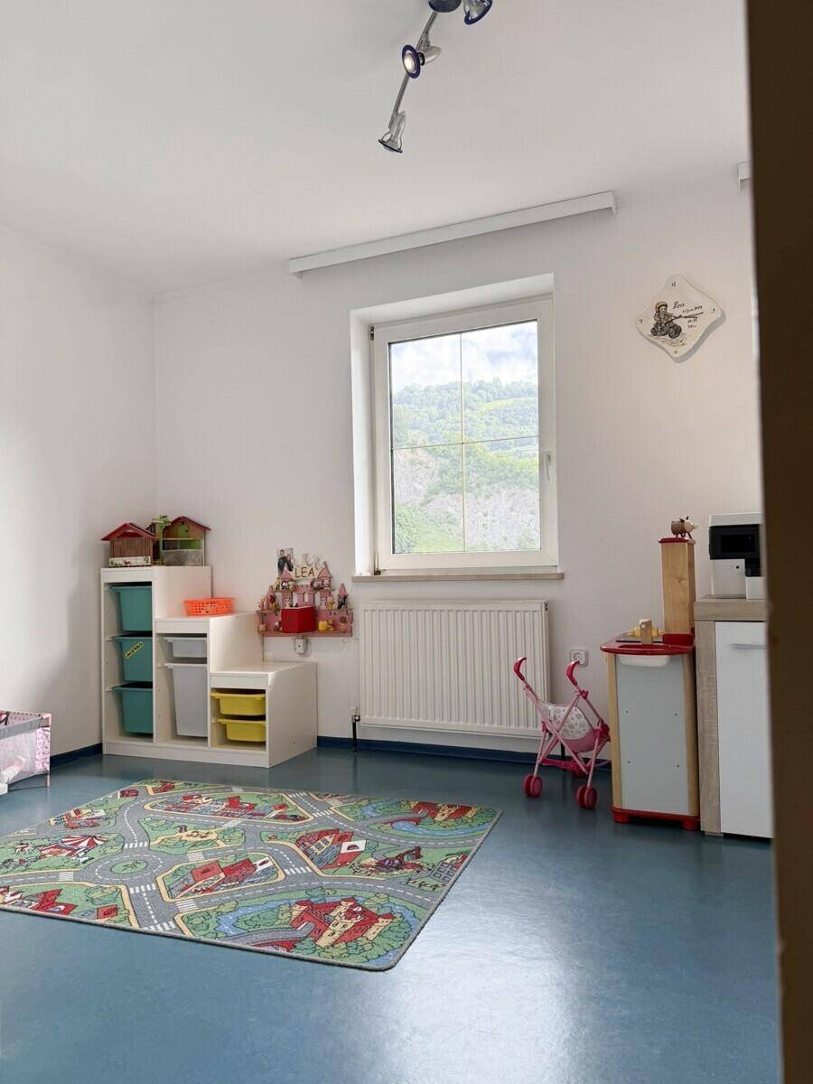 Helles Kinderzimmer mit blauem Boden, Spielteppich und Fenster mit Blick ins Grüne.