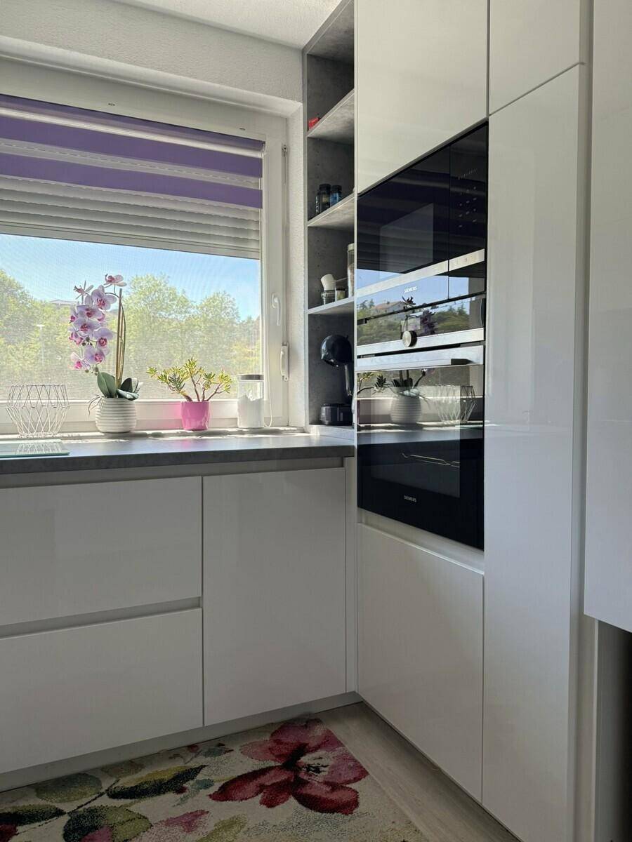 Moderne Küche mit weißen Hochglanzfronten, Einbauofen und Fenster mit Blick ins Freie.