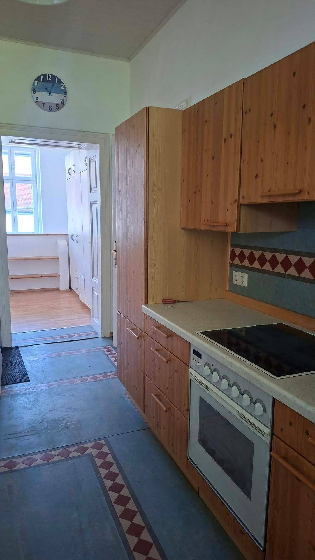 Küche mit Holzschränken, Elektroherd und gemusterten Fliesenböden, angrenzend an einen weiteren Raum.