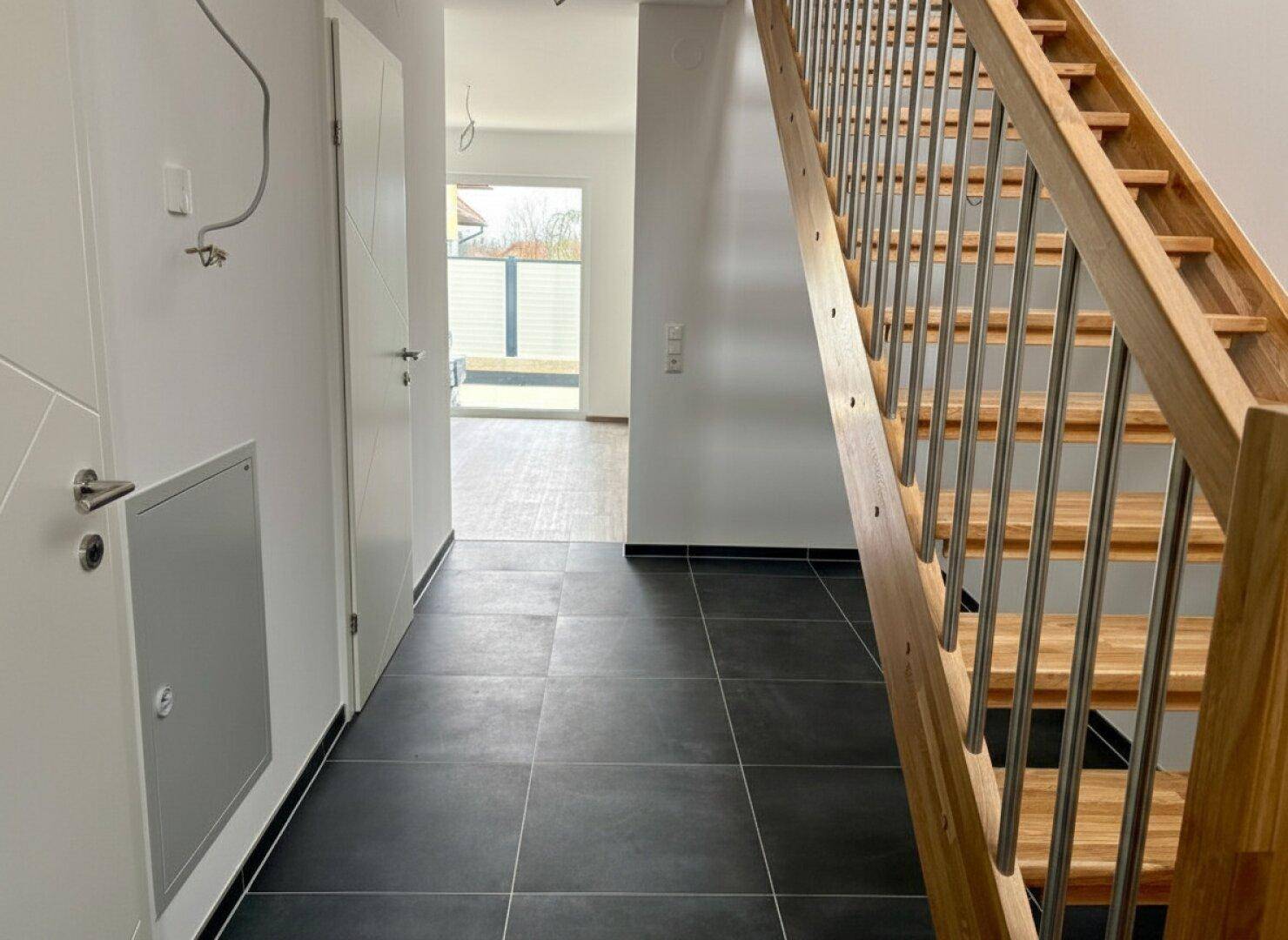 Eingangsbereich mit moderner Holztreppe, dunklen Fliesen und Zugang zu weiteren Räumen.