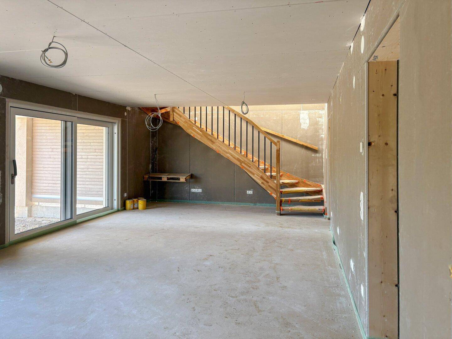 Offener Wohnbereich im Rohbau mit Holztreppe und viel Tageslicht.