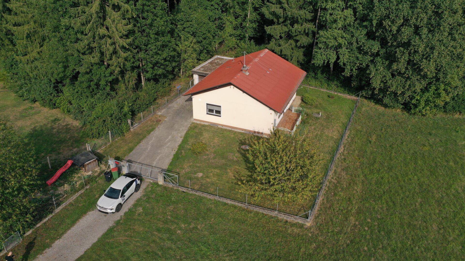 Luftaufnahme eines Einfamilienhauses mit rotem Dach, umgeben von einem eingezäunten Garten und Bäumen.
