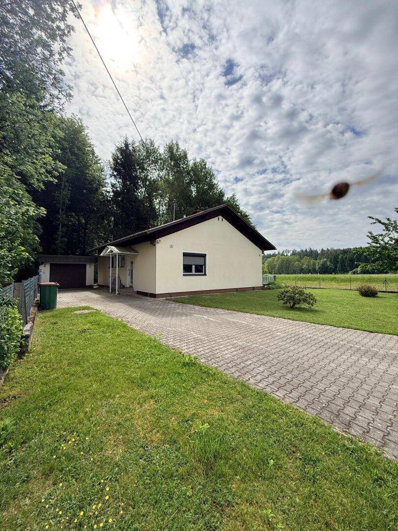 Vorderansicht eines Einfamilienhauses mit gepflasterter Auffahrt, Rasenfläche und umgebenden Bäumen unter bewölktem Himmel.