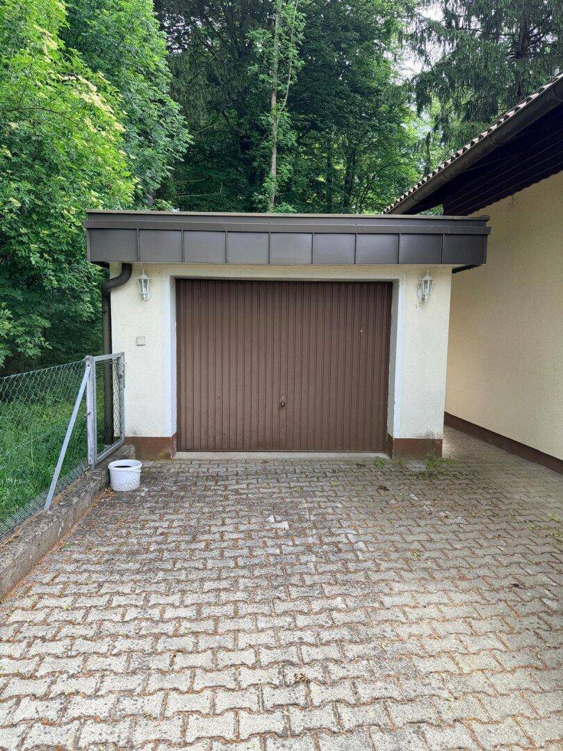 Einzelgarage mit braunem Tor und gepflastertem Vorplatz, umgeben von Bäumen.