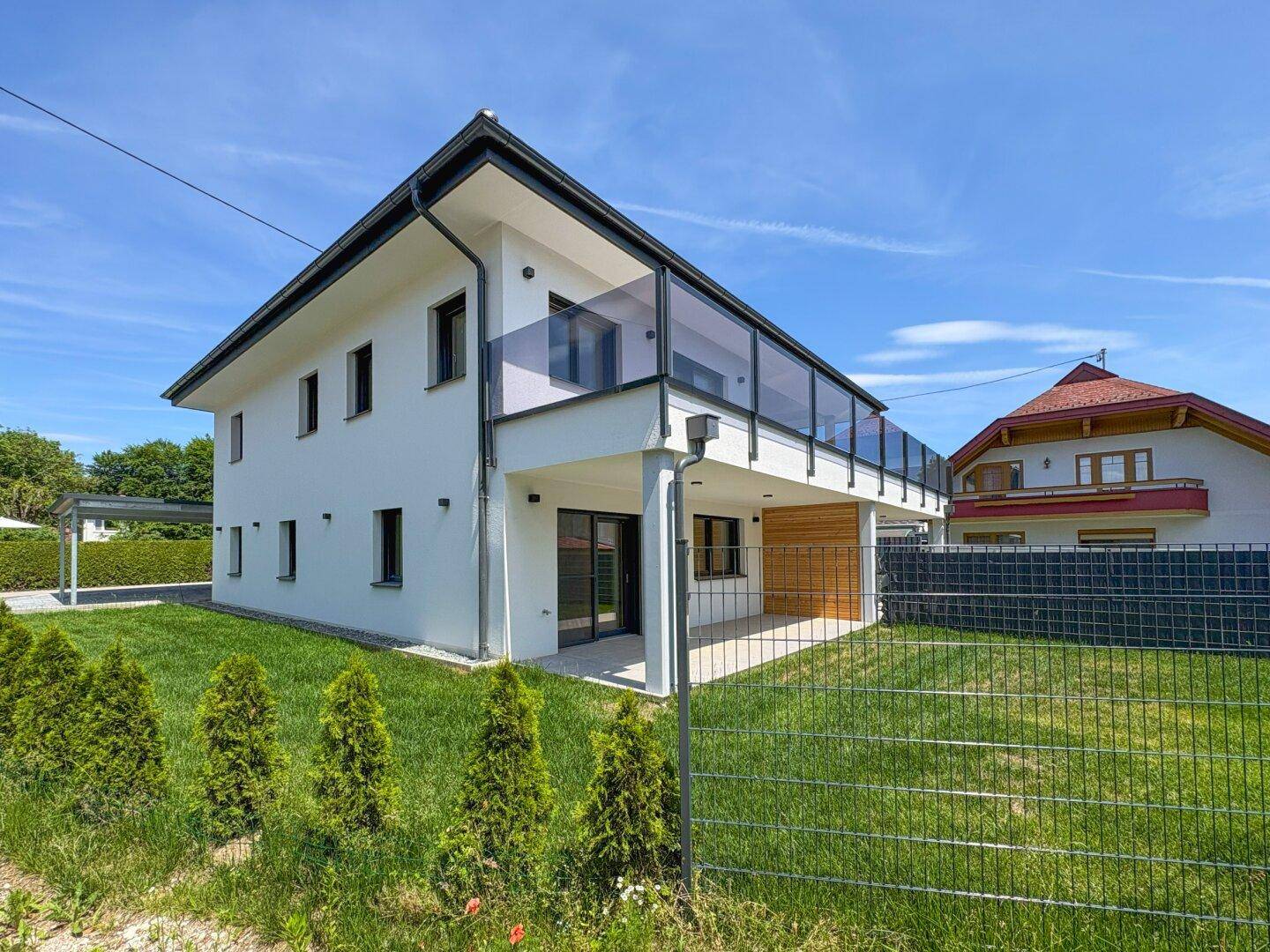 Modernes Einfamilienhaus mit weißer Fassade, Balkon und gepflegtem Gartenbereich unter blauem Himmel.