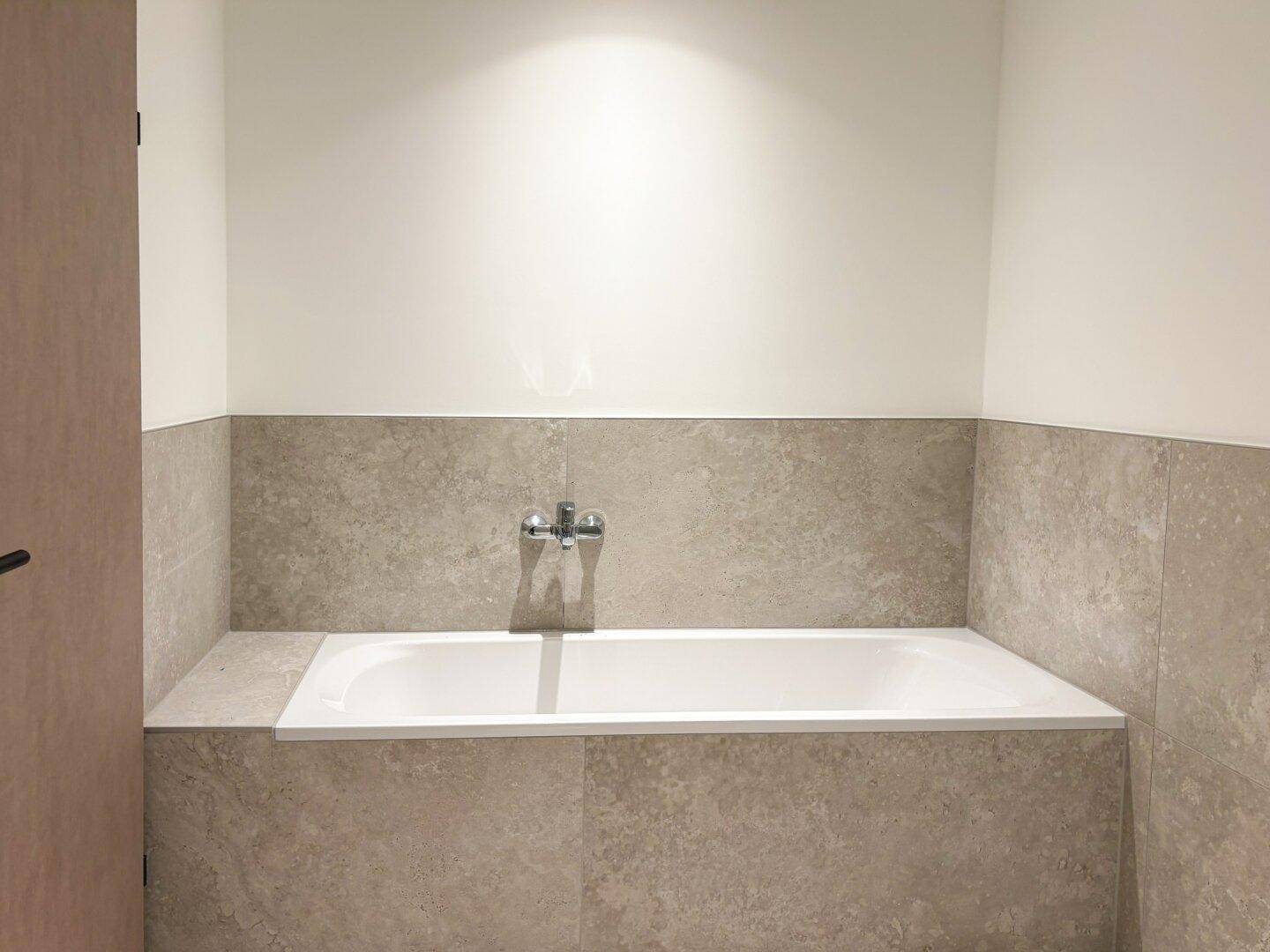 Helles Badezimmer mit eingebauter Badewanne und modernen grauen Fliesen an den Wänden.