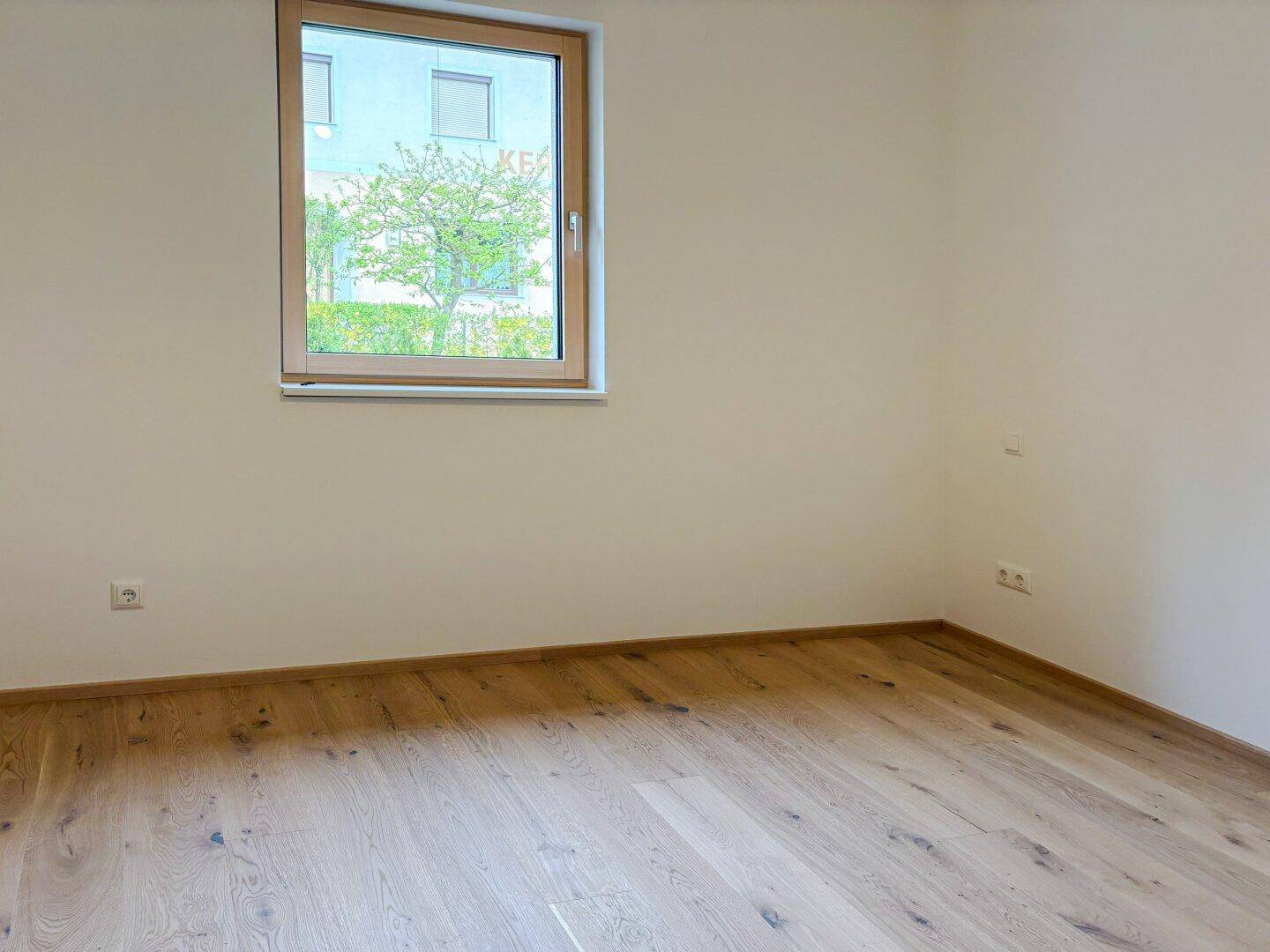 Leeres Schlafzimmer mit hellem Holzfußboden und einem Fenster mit Blick ins Grüne.