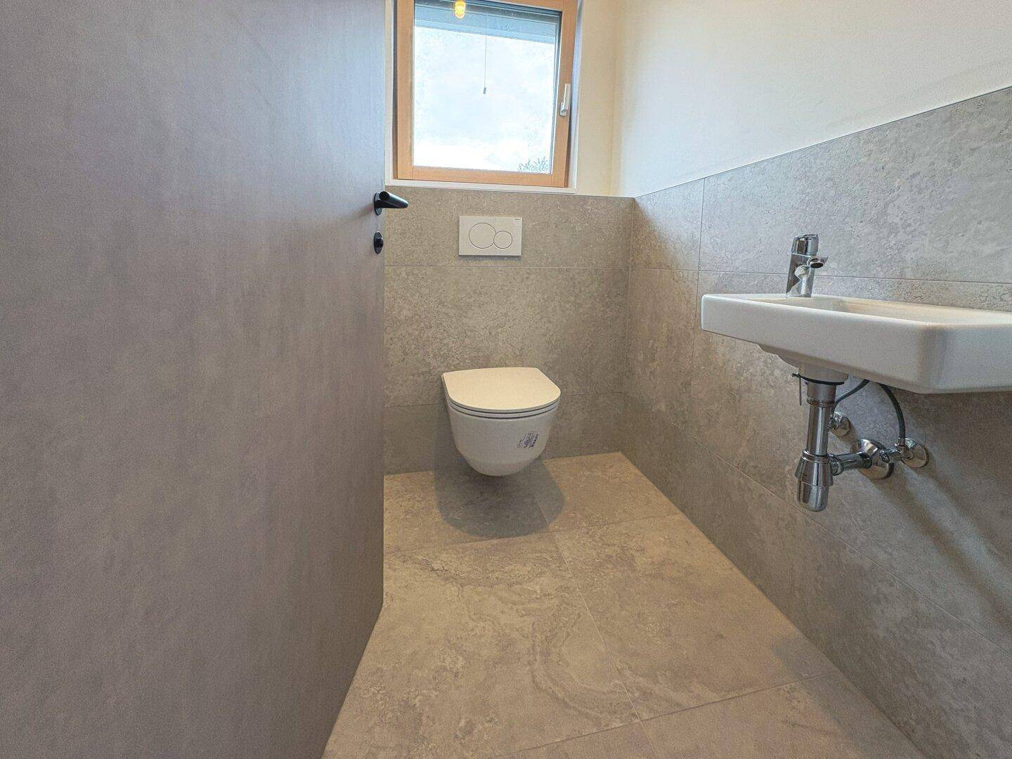 Gäste-WC mit wandhängender Toilette, kleinem Waschbecken und Fenster für gute Belüftung.