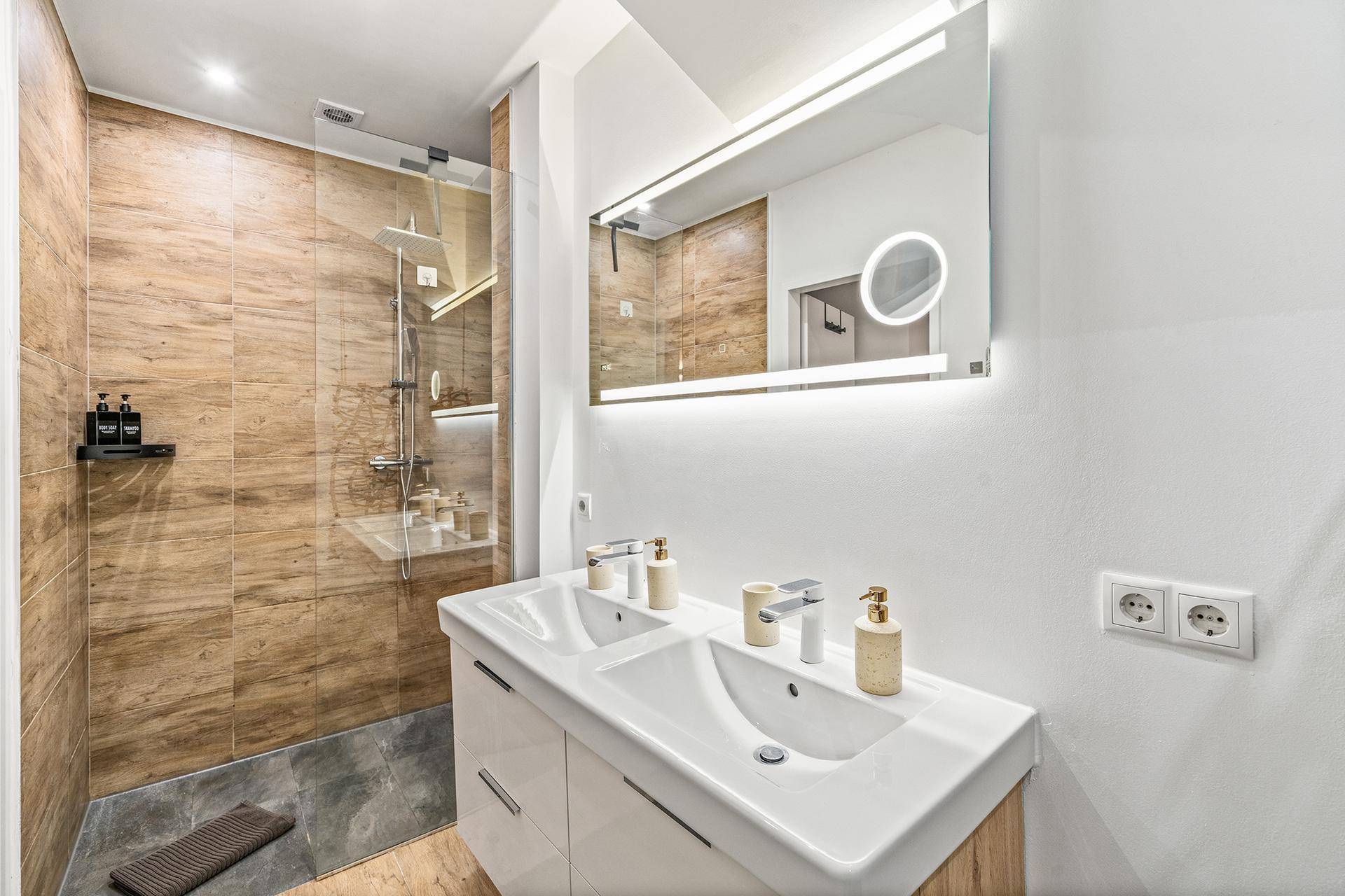 Modernes Badezimmer mit Doppelwaschbecken, beleuchtetem Spiegel und begehbarer Dusche.