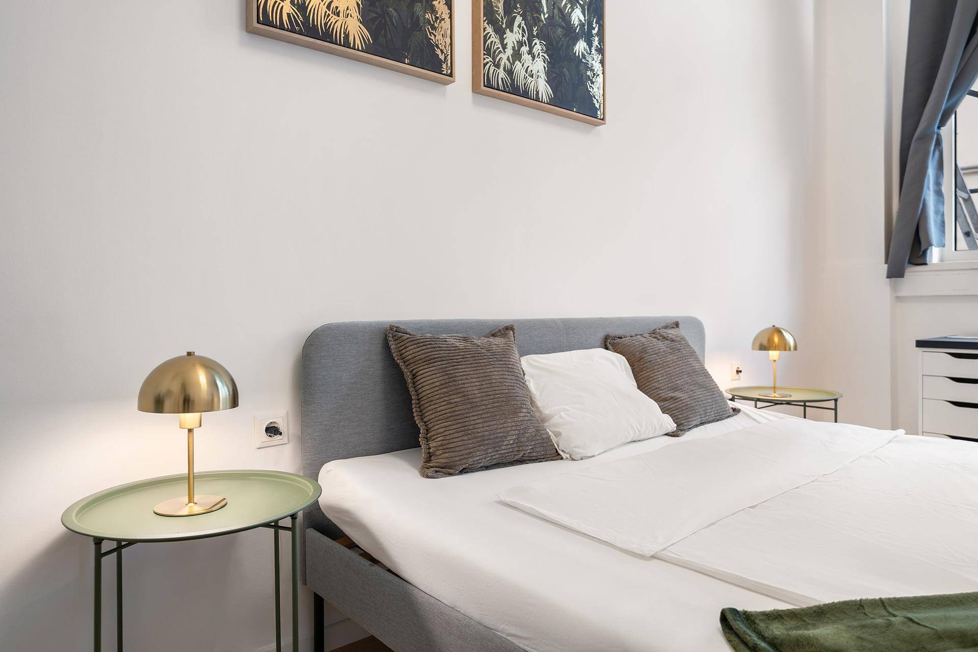 Detailansicht des Bettes mit Nachttisch und goldener Lampe in einem hellen Schlafzimmer.
