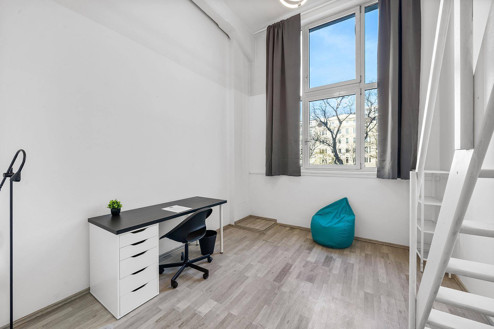 Arbeitsbereich mit Schreibtisch und Stuhl unter einem großen Fenster in einem hellen Raum.