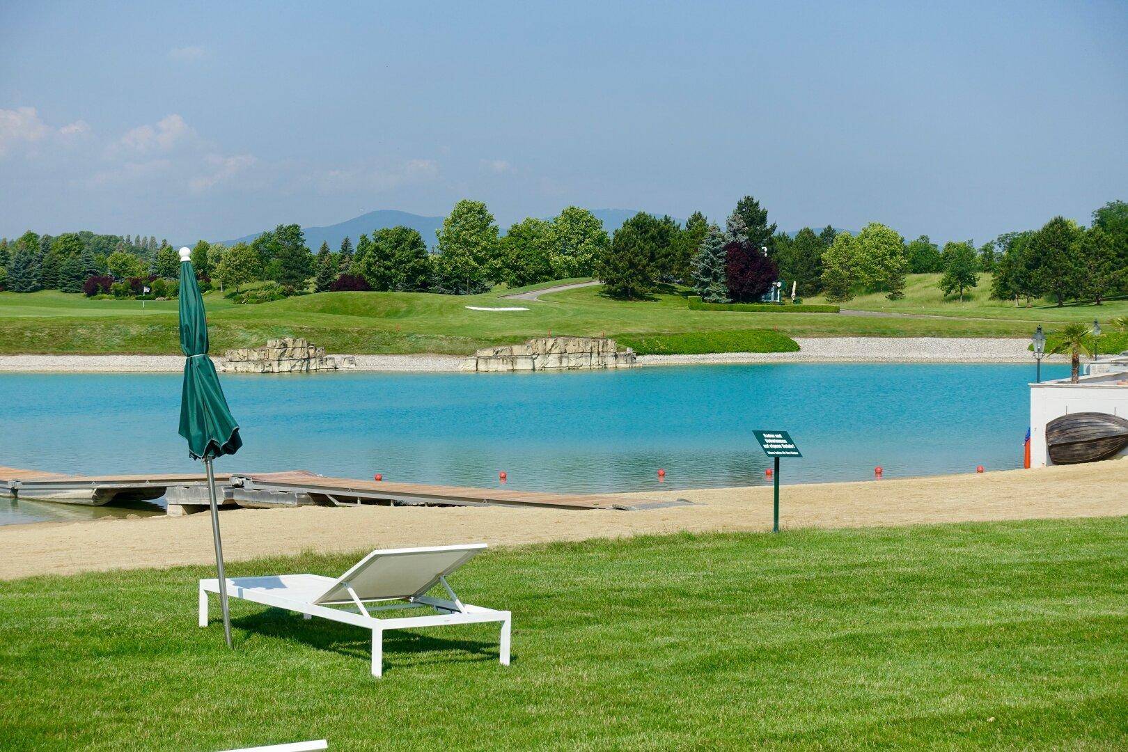 Idyllische Seelandschaft mit grüner Rasenfläche, einem Liegestuhl und Golfplatz im Hintergrund.