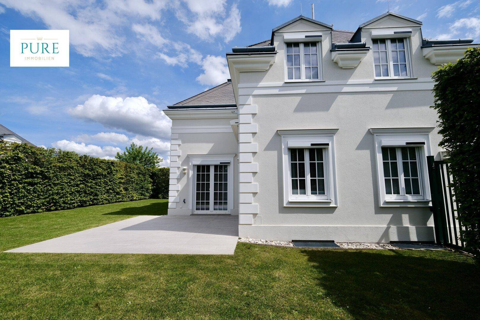 Seitenansicht des Hauses mit Terrasse, gepflegtem Rasen und grüner Hecke.
