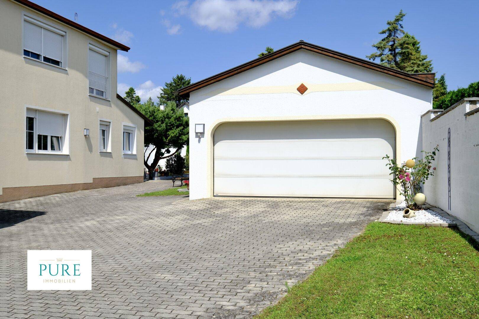 Freistehende Garage mit weißem Rolltor und gepflasterter Zufahrt neben einem Wohnhaus.