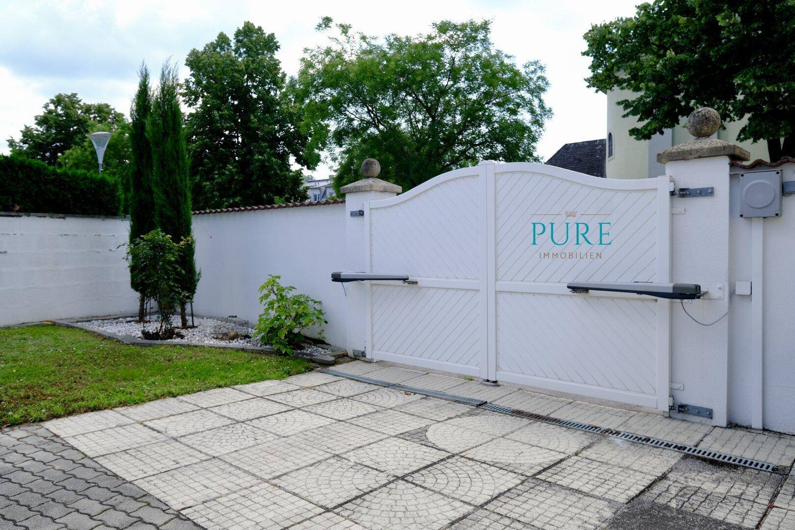 Weißes automatisches Einfahrtstor mit gepflastertem Bereich und einer hohen weißen Mauer.