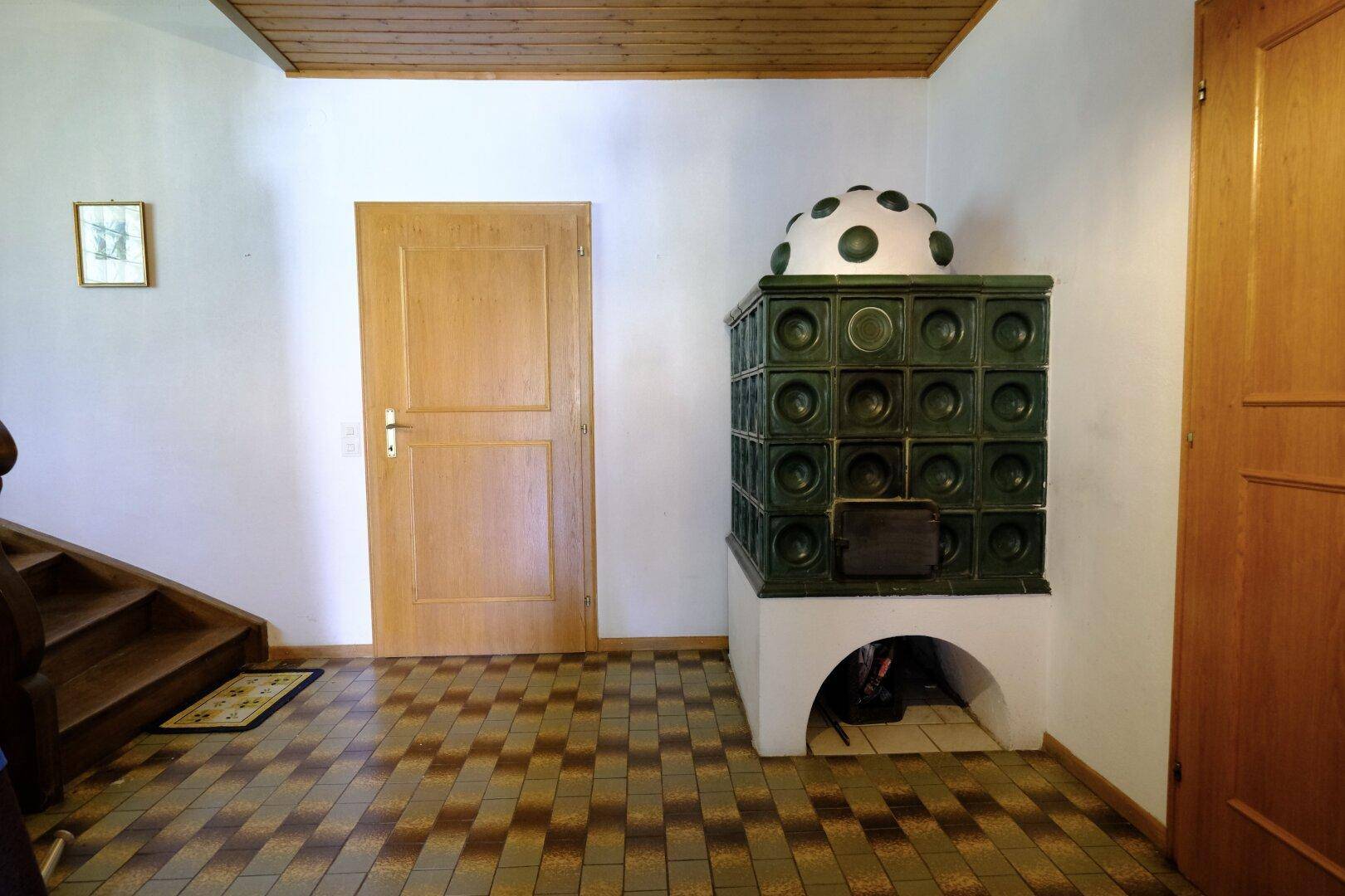 Bereich mit grünem Kachelofen, Fliesenboden und Holzdecke neben einer Treppe.