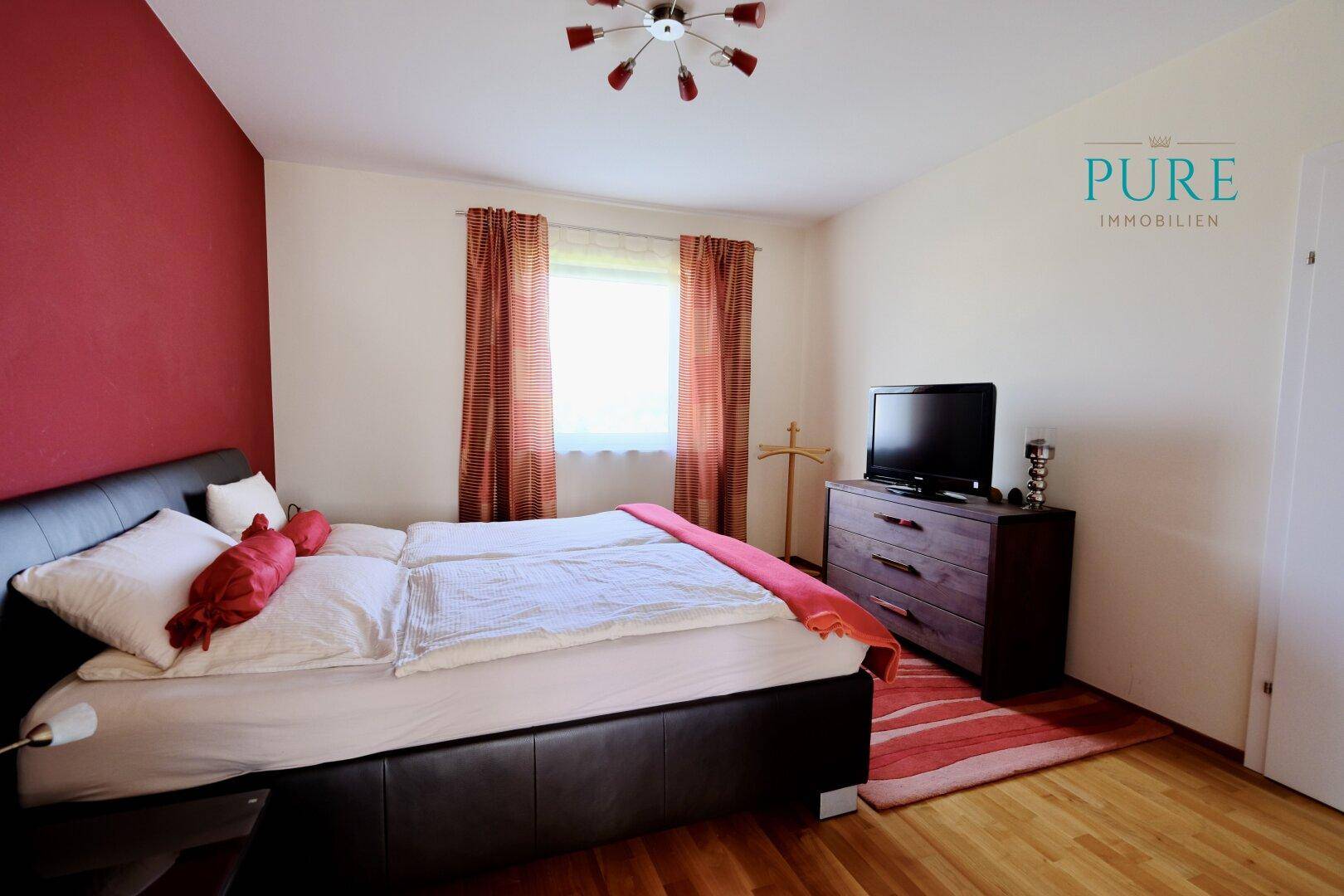 Gemütliches Schlafzimmer mit Doppelbett, Holzboden und roter Akzentwand, hell und einladend.