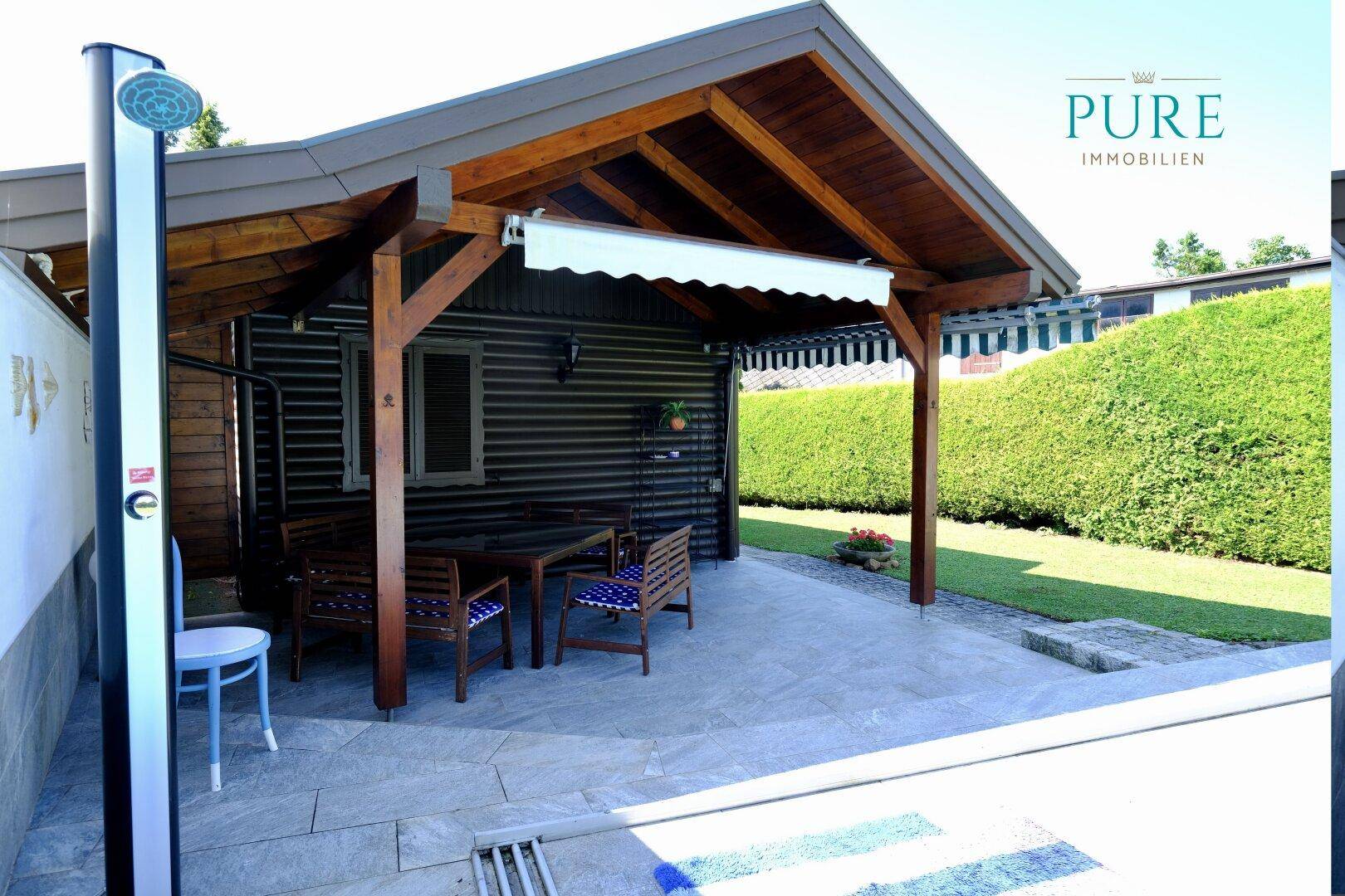 Überdachte Terrasse mit Sitzgelegenheiten und Außendusche neben dem Poolbereich.