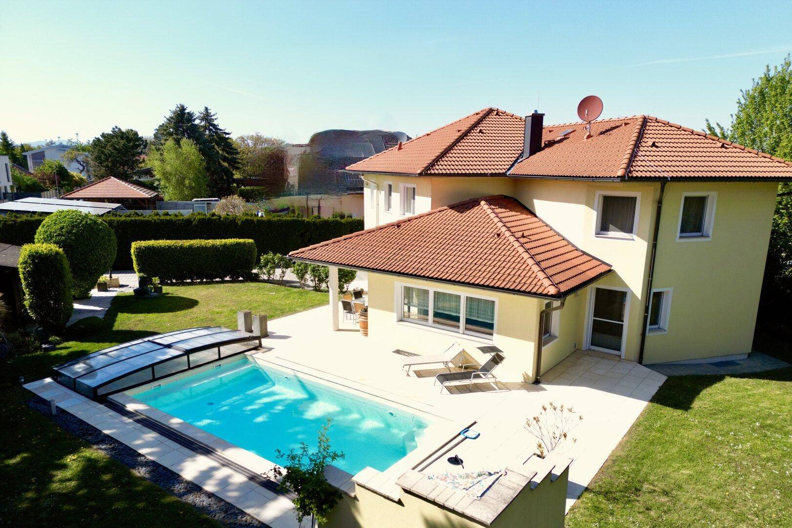 Weitläufiger Gartenbereich mit großem Pool und sonniger Terrasse, umgeben von grünen Hecken.