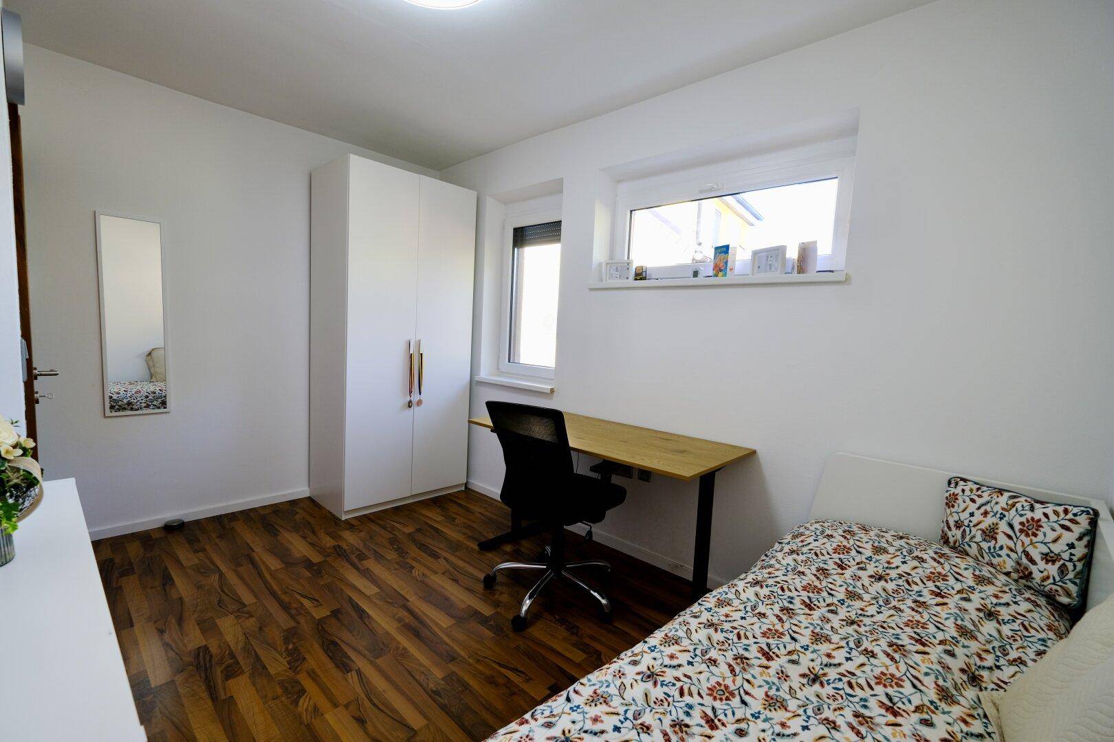 Freundliches Schlafzimmer mit Holzboden, Schreibtisch und Kleiderschrank, hell und funktional eingerichtet.