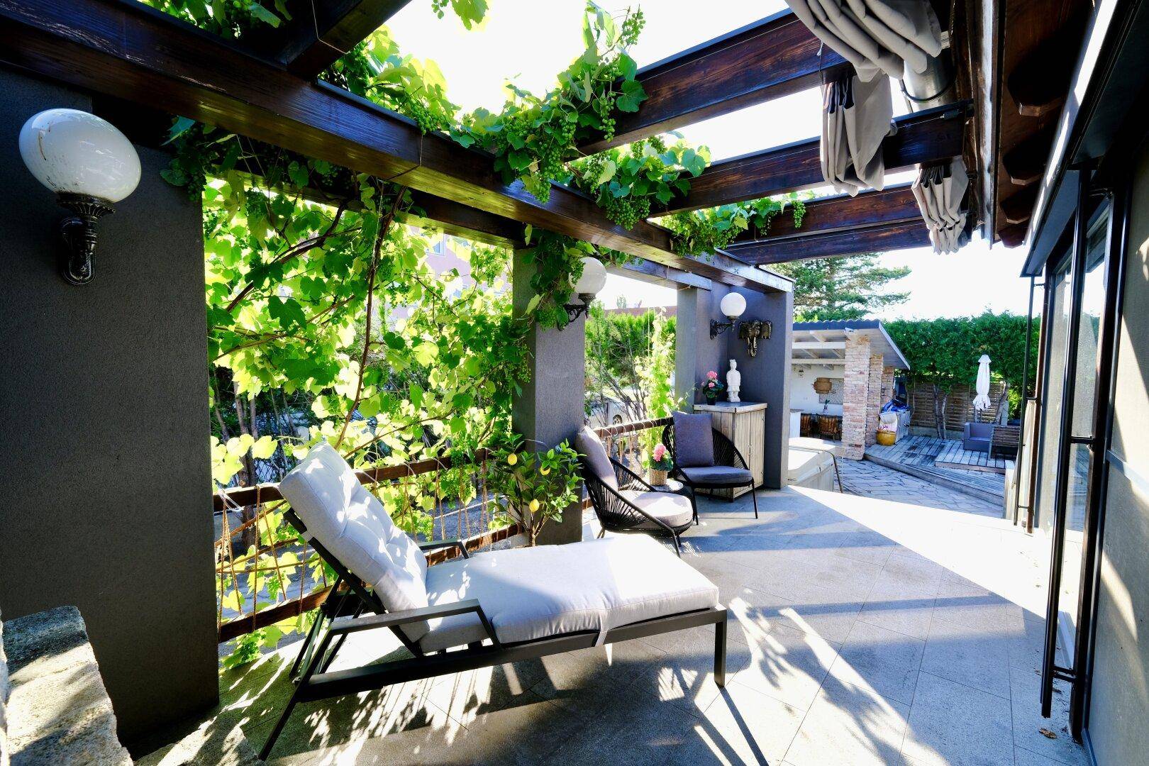 Überdachte Terrasse mit bequemen Liegen und Sitzgelegenheiten, umgeben von üppigem Grün.
