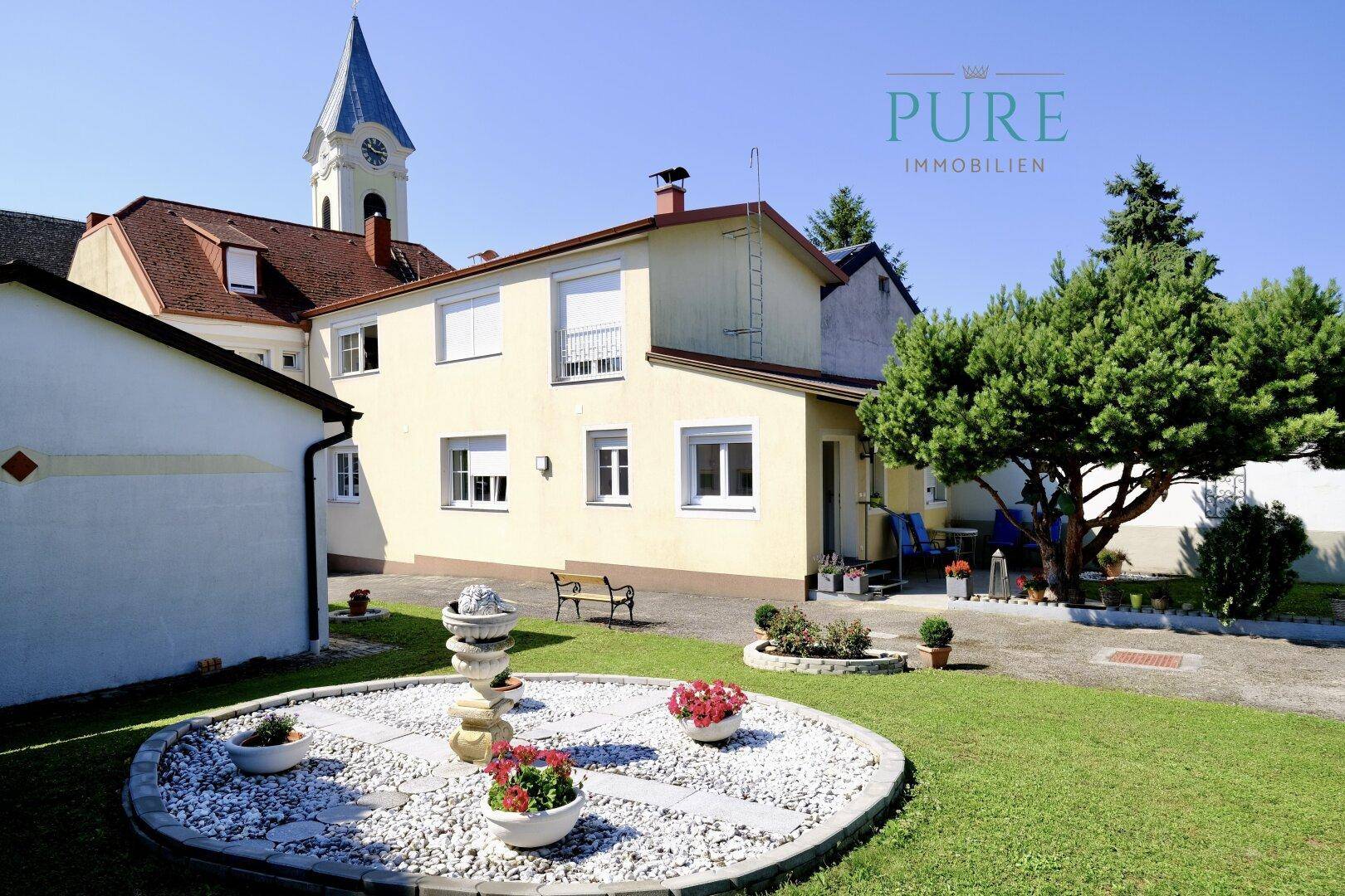 Gelbes Haus mit gepflegtem Vorgarten und einem Kirchturm im Hintergrund unter blauem Himmel.