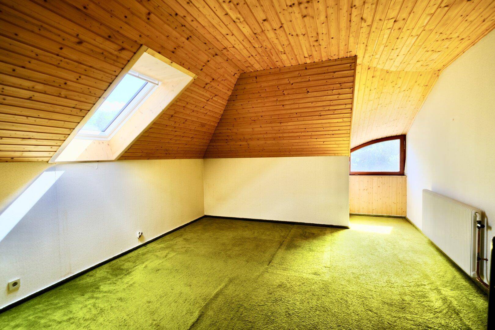 Dachzimmer mit schräger Holzdecke, grünem Teppichboden und einem Dachfenster für Tageslicht.