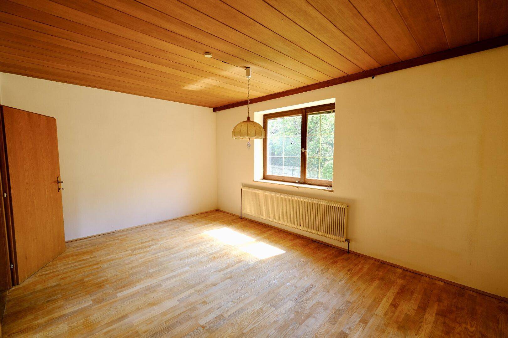 Leerer Raum mit Holzdeckenverkleidung, Parkettboden und Fenster mit Blick ins Grüne.