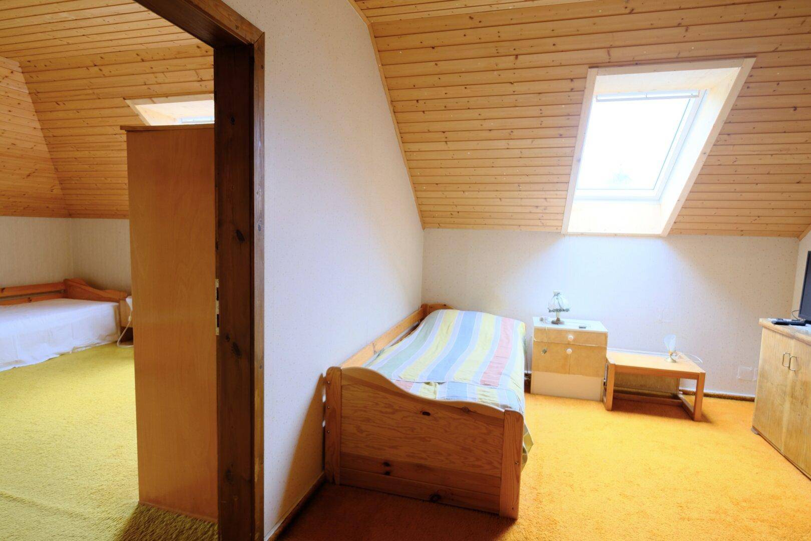 Gemütliches Dachschlafzimmer mit Holzdecke, gelbem Teppich und viel natürlichem Licht durch das Dachfenster.
