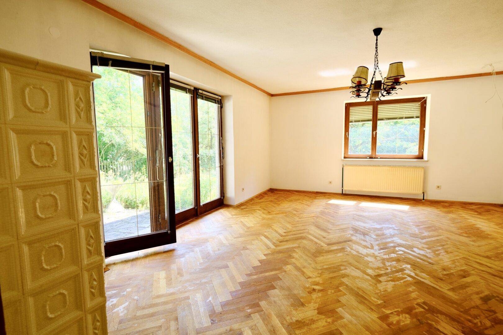 Helles Wohnzimmer mit traditionellem Kachelofen, Fischgrätparkett und Zugang zur Terrasse mit Grünblick.