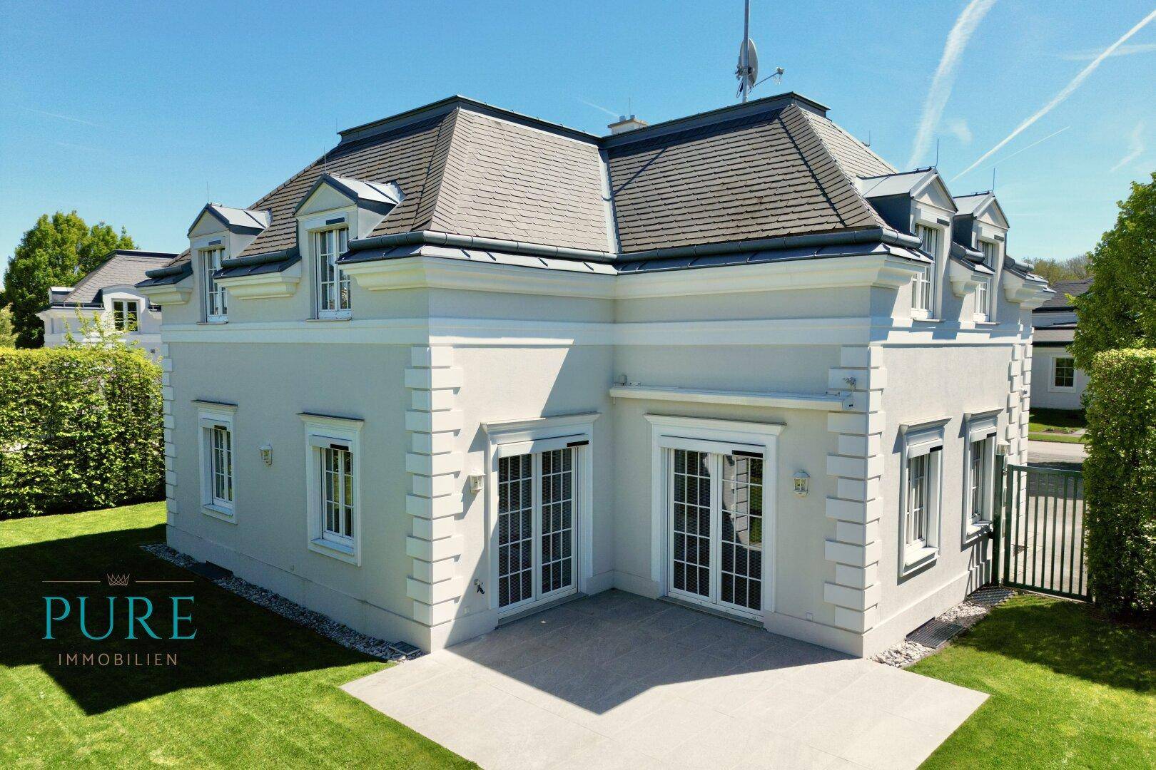 Elegantes Einfamilienhaus mit Mansarddach, heller Fassade und einer gepflasterten Terrasse zum Garten.