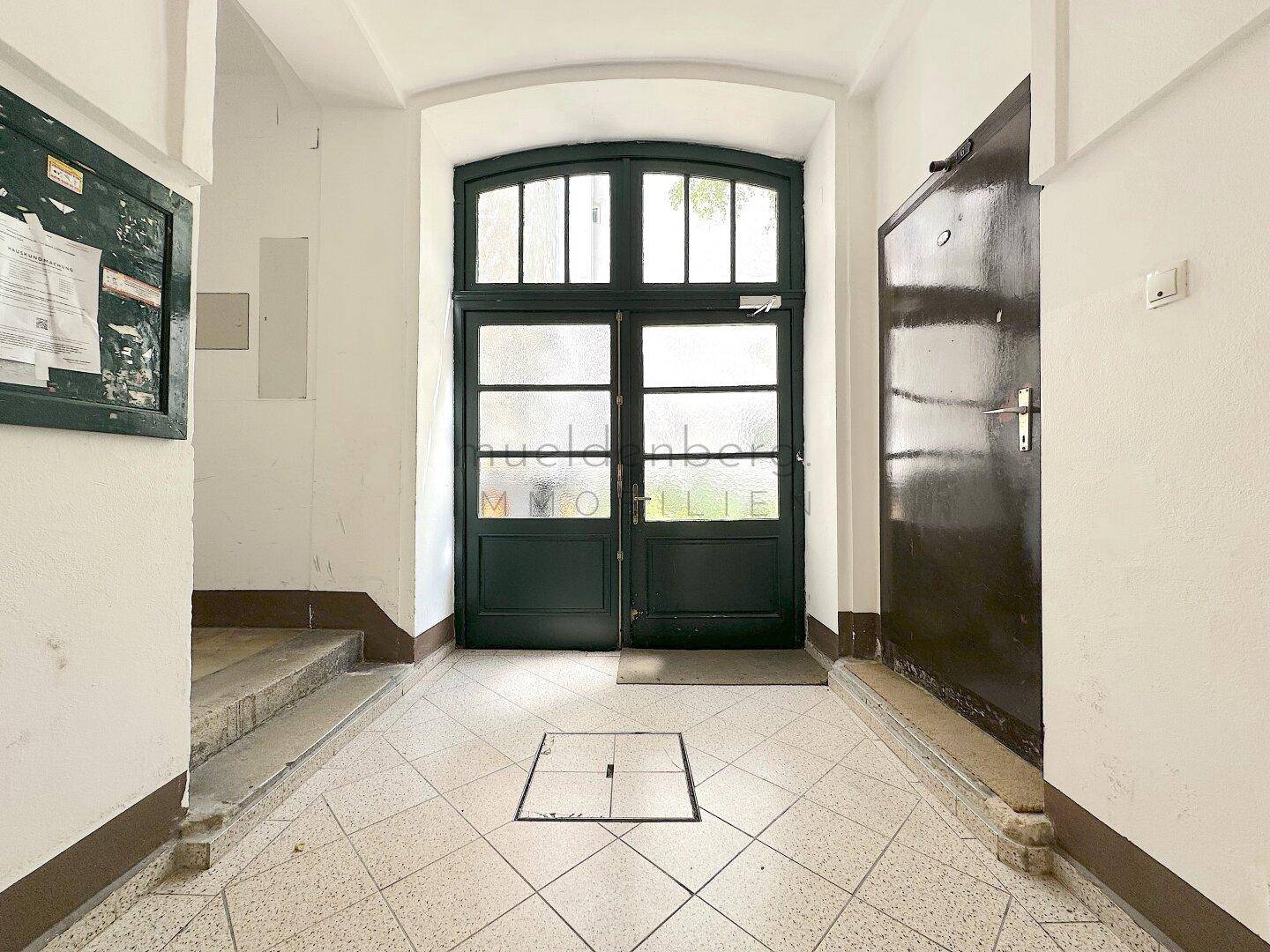 Eingangsbereich eines Altbaus mit doppelter Holztür und Fliesenboden, der in den Hausflur führt.