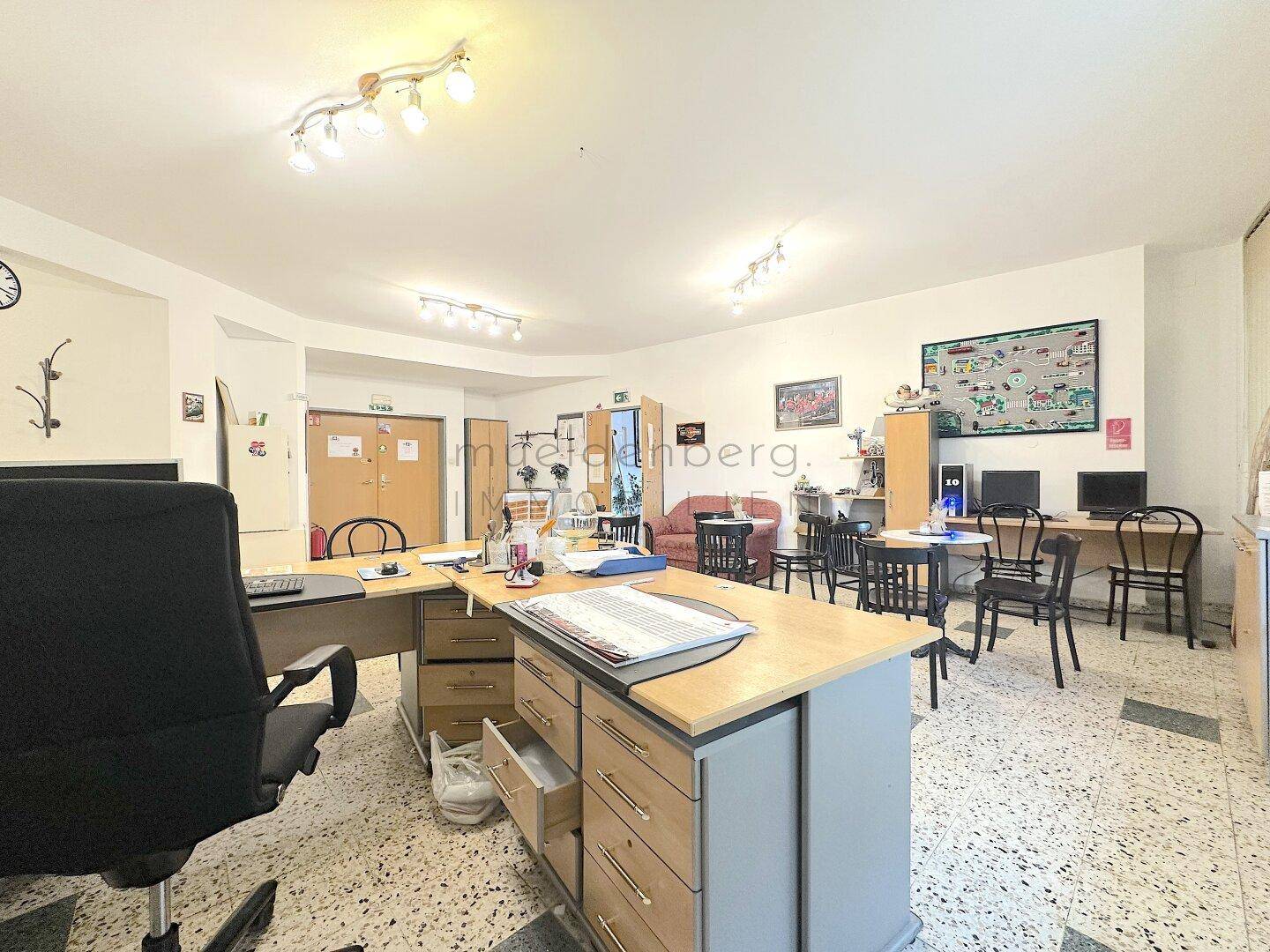 Büroarbeitsplatz mit großem Schreibtisch, Bürostuhl und Blick auf den offenen Bürobereich mit Sitzgruppen.