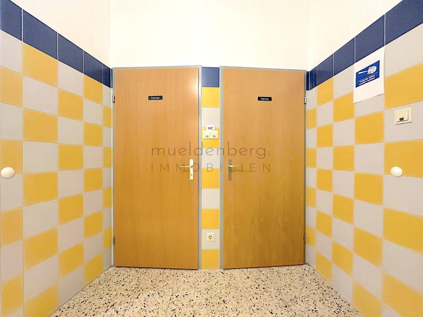 Eingang zu den Toiletten mit zwei Holztüren und farbenfrohen gelb-blauen Fliesen an den Wänden.