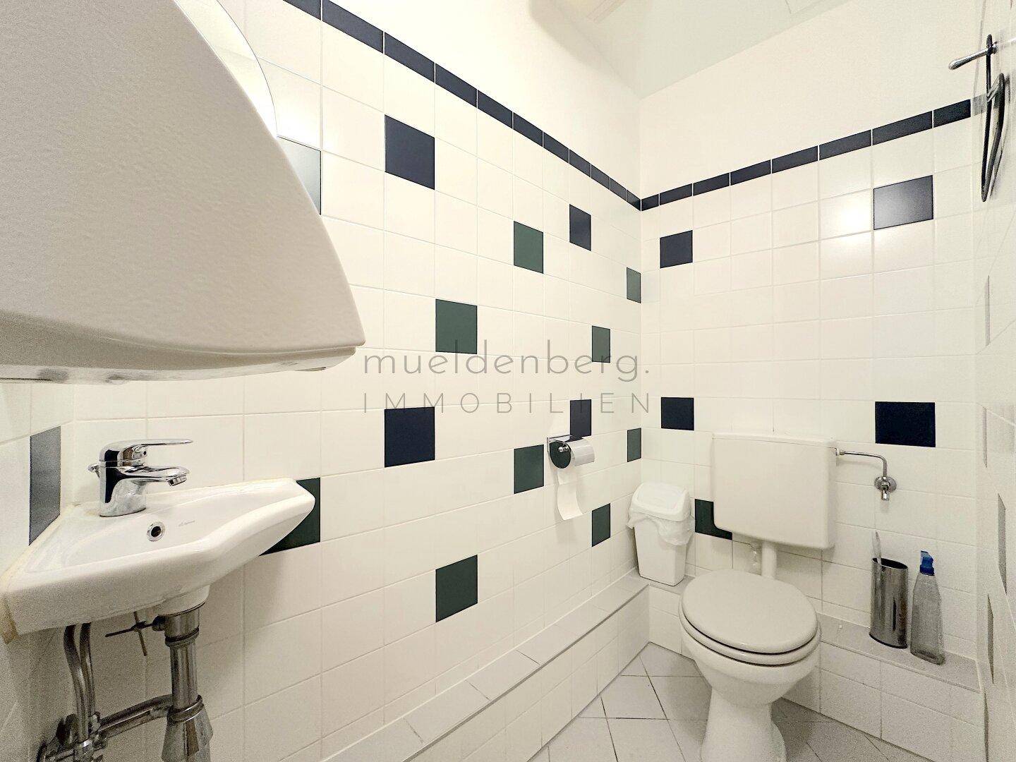 Heller Toilettenraum mit weiß-dunkelgrün gefliesten Wänden, einem Waschbecken und einer Toilette.