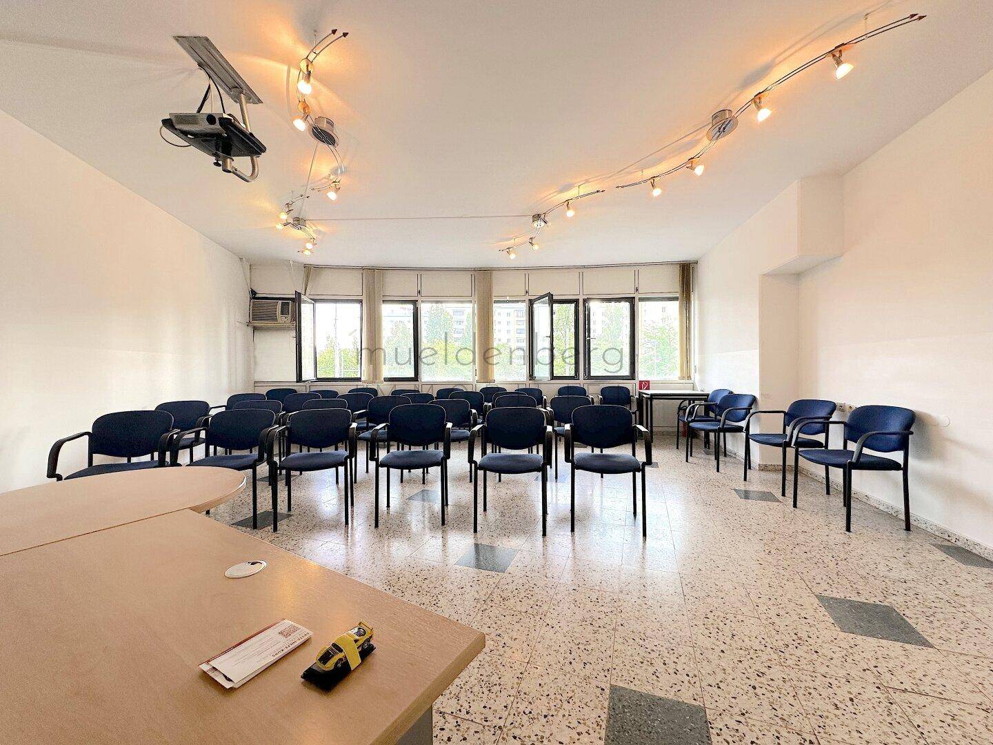 Heller Konferenzraum mit vielen Fenstern, blauen Stühlen und einem Projektor, der eine gute Sicht bietet.
