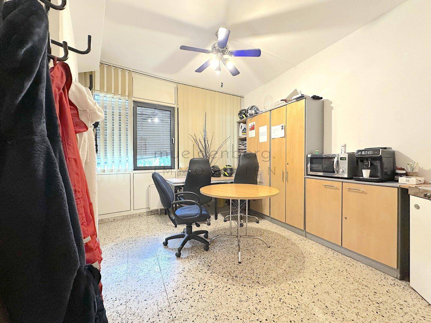 Geräumiges Büro mit funktionaler Einrichtung, integrierter Küchenzeile und Deckenventilator, ideal für den Arbeitsalltag.
