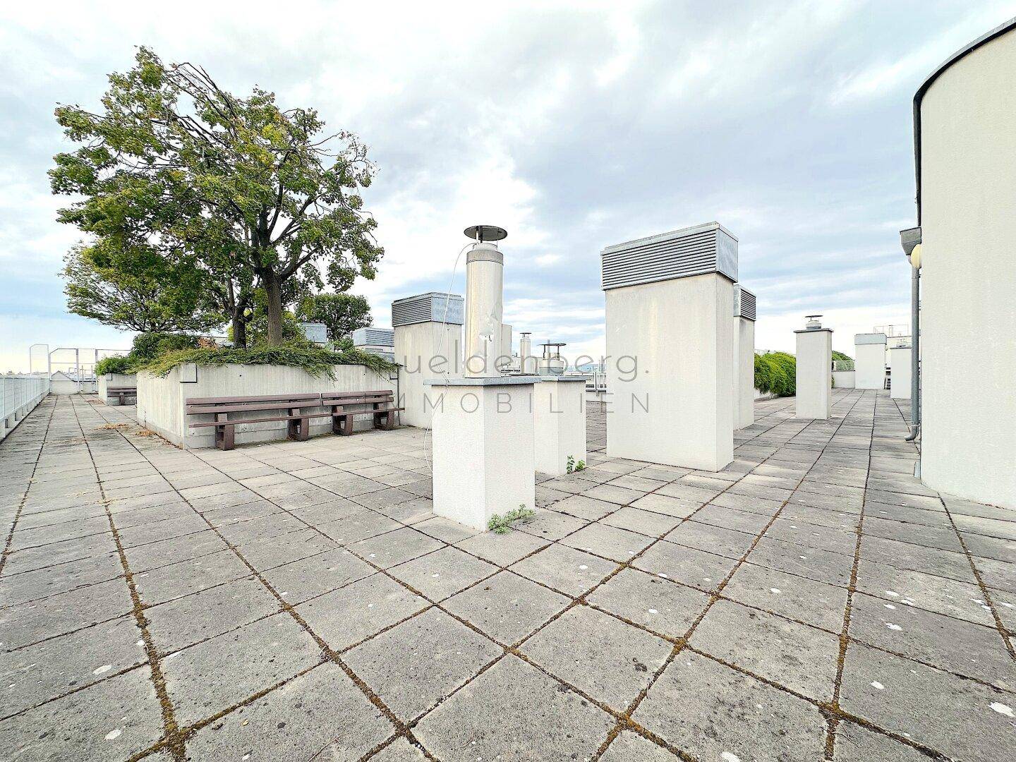 Weitläufige Dachterrasse mit Betonplatten, Sitzbänken und einem Baum, umgeben von technischen Installationen.