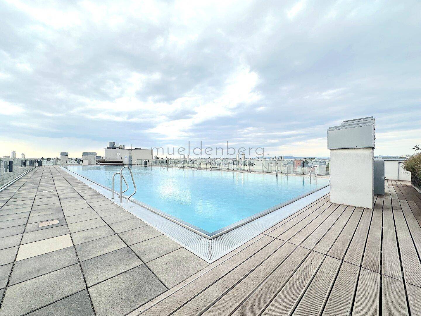 Dachpool mit Holz- und Fliesenbelag, umgeben von einer städtischen Skyline unter einem bewölkten Himmel.