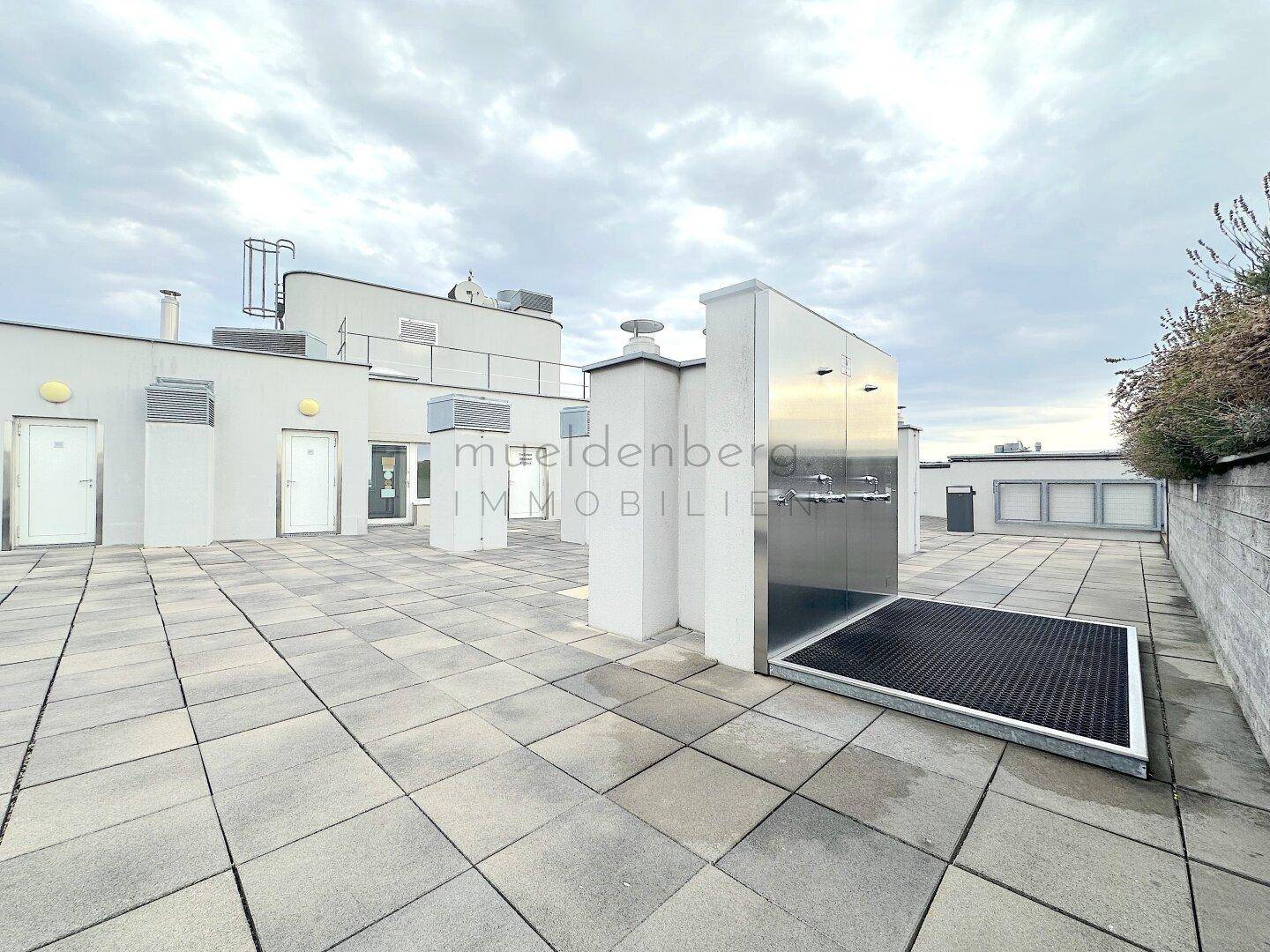 Dachbereich mit Betonplatten, weißen Gebäudestrukturen und einer Außendusche unter einem bewölkten Himmel.