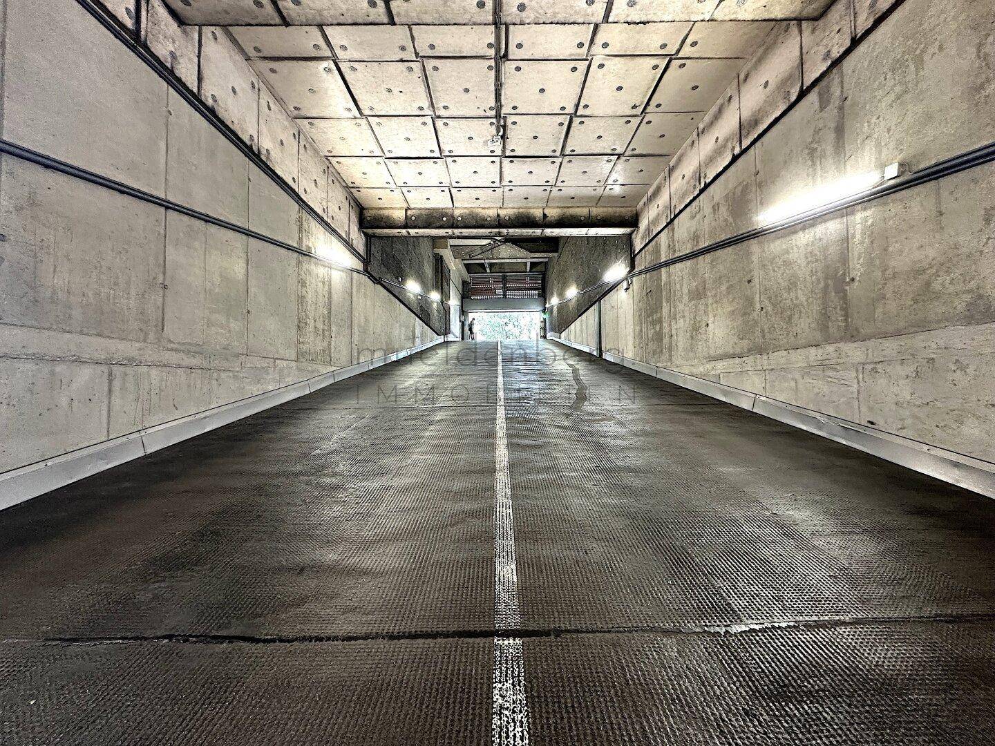 Lange Rampe führt hinab in die Tiefgarage, umgeben von Betonwänden und -decken, gut beleuchtet.