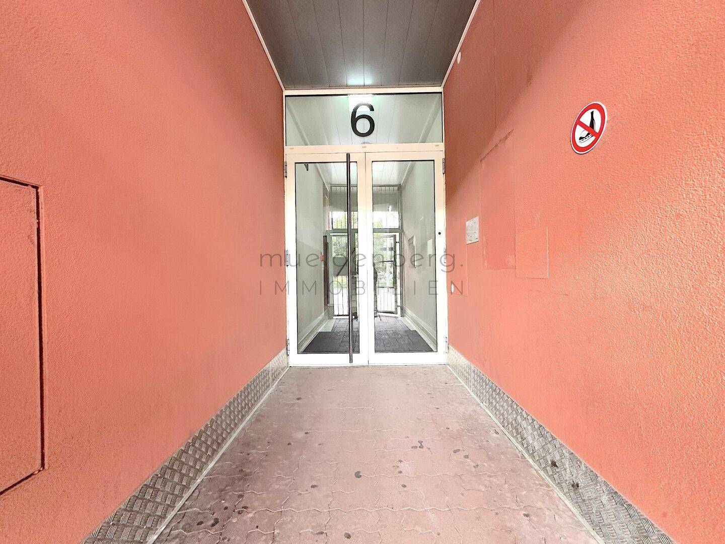 Eingangskorridor mit Rampe führt zu Glastüren mit der Nummer 6, umgeben von roten Wänden.