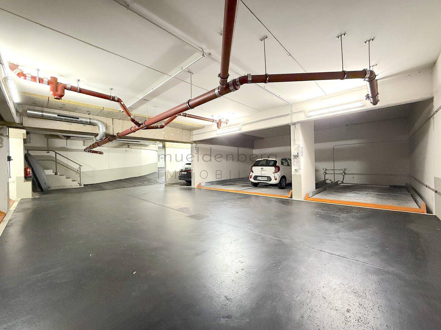 Helle Tiefgarage mit mehreren Stellplätzen und sichtbaren Installationen an der Decke.