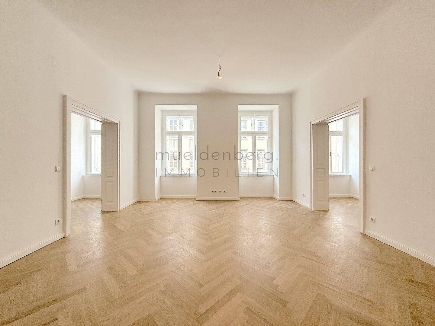 Geräumiges, helles Zimmer mit Fischgrätparkett und zwei großen Fenstern, ideal als Wohnzimmer.